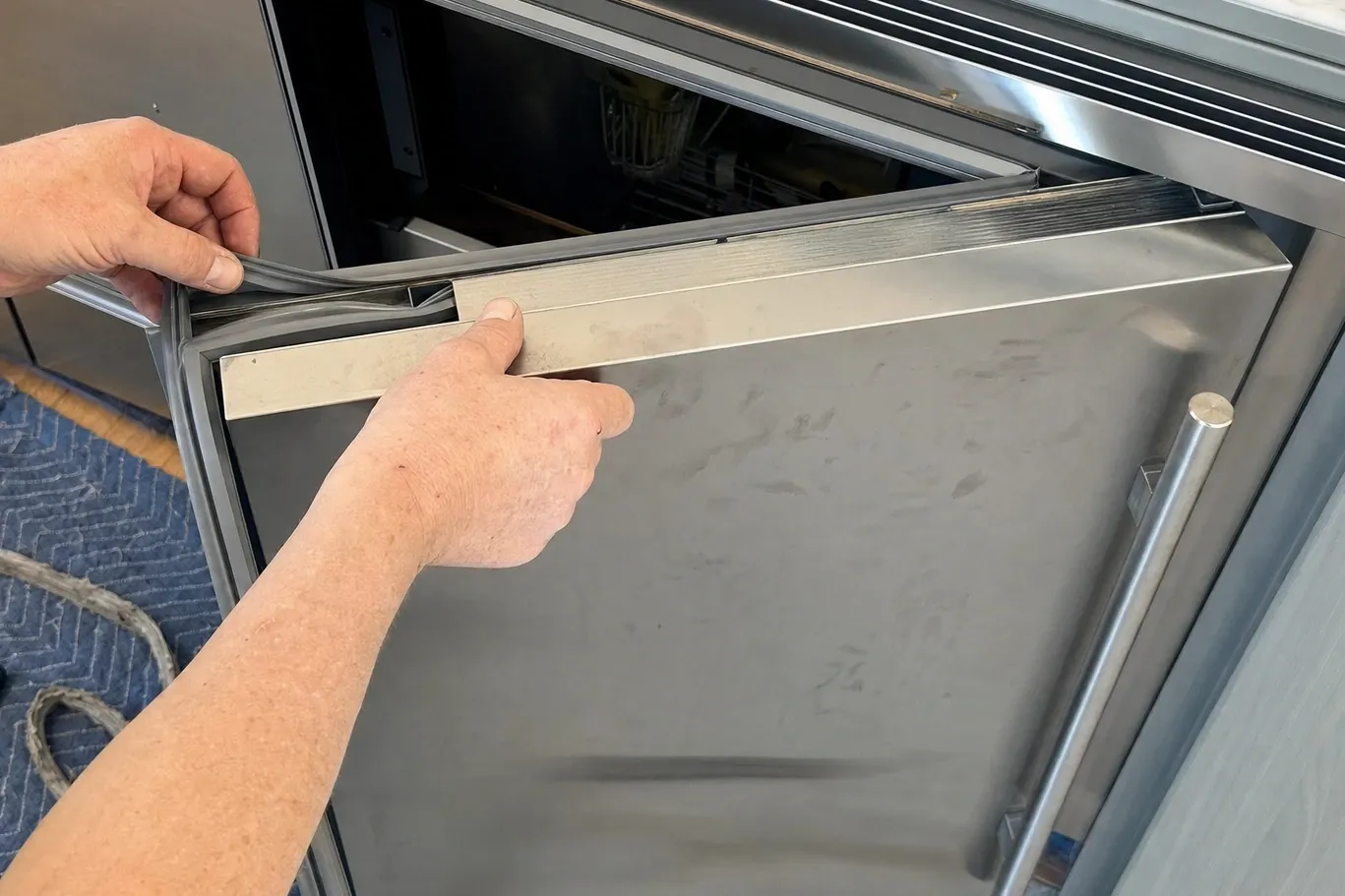 Two hands pulling the flexible door gasket loose from the top edge of a stainless door, a wire basket visible inside