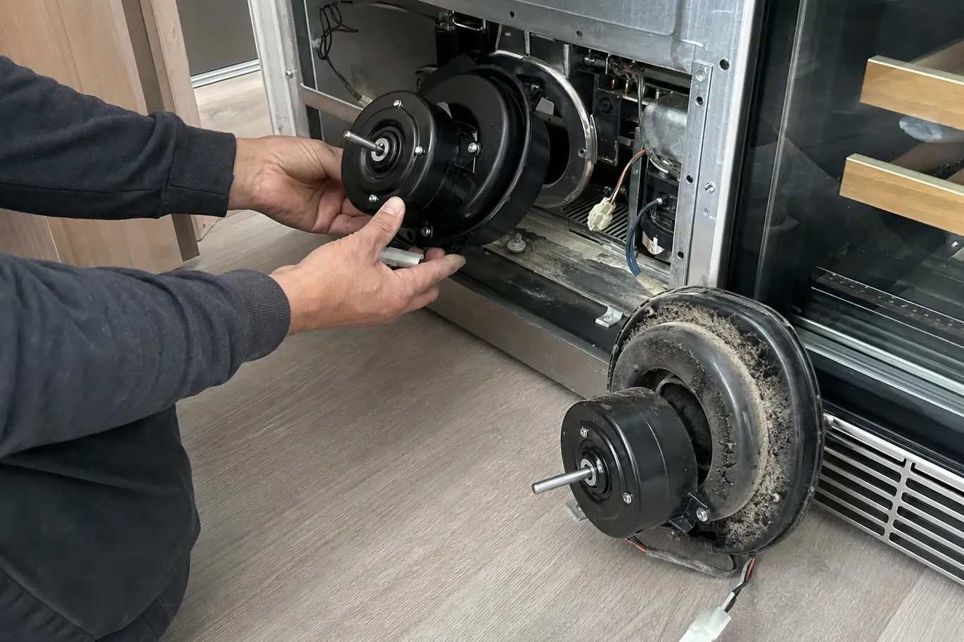 Hand holding a new condenser fan motor at an open machine compartment, the old motor and its dust-caked shroud on the floor