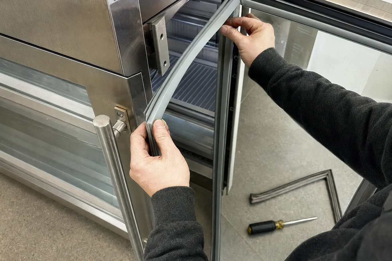 Hands seating a new grey gasket into the door channel of a stainless glass-door unit, old gasket and screwdriver on the floor