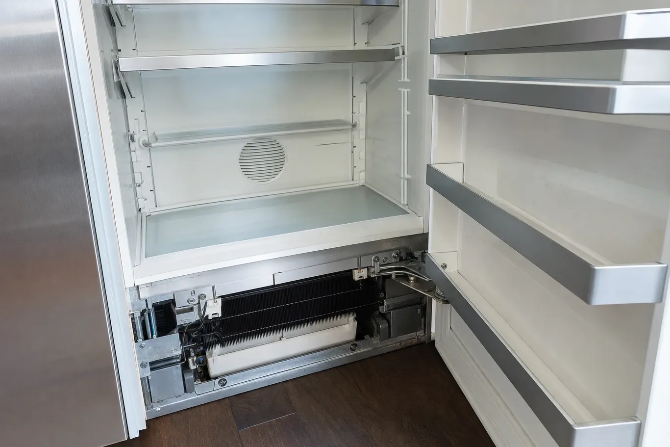 Empty refrigerator interior with the lower access panel removed, exposing the finned evaporator coil and the drain pan beneath the shelves